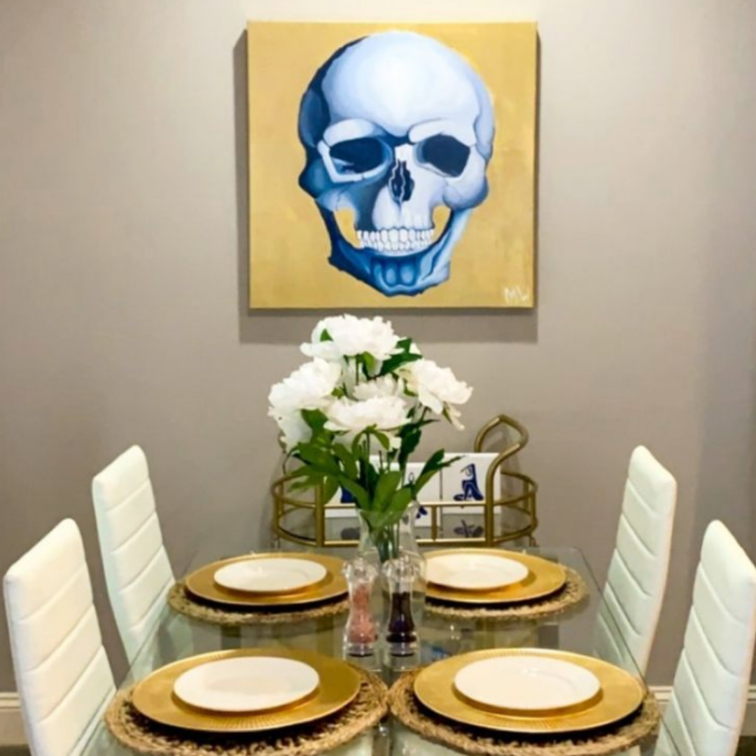 A large canvas hangs above a modern dining room table set with white and gold plates and tall white chairs around it. The painting features a large blue skull on a shiny gold background.