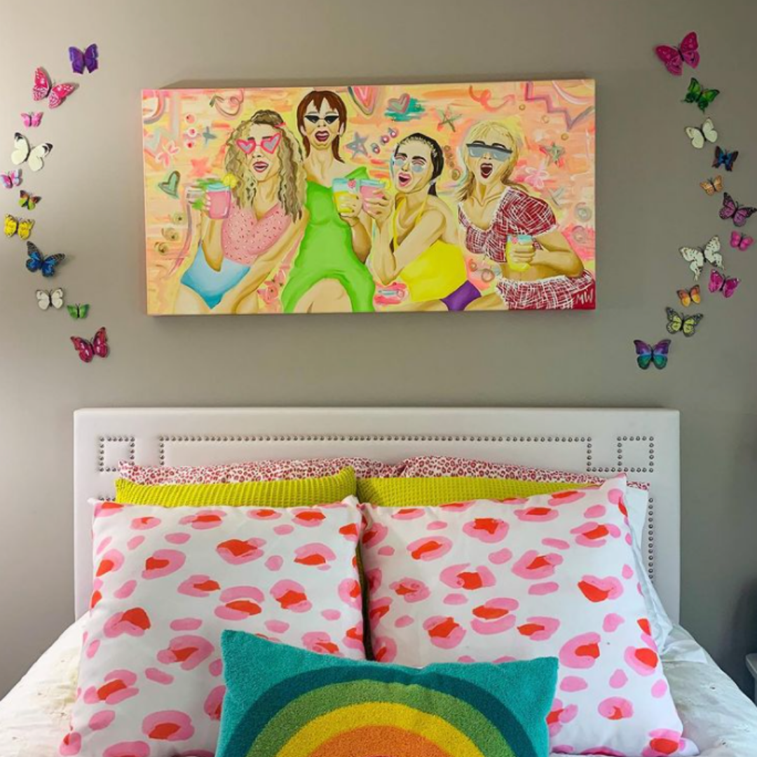 A bright and colorful canvas painting of the characters from Sex in the City with drinks in their hands hangs on a wall surrounded by cut out rainbow butterflies above a bed made with pink and rainbow pillows.