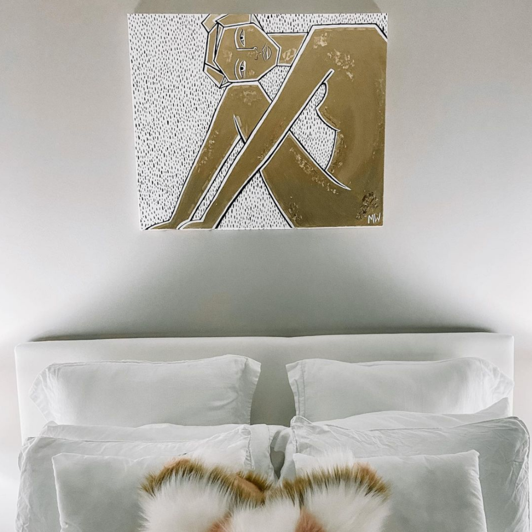 A canvas painting with a white background and black polka dots hangs above a white bed with fluffy pillows. On the canvas is a women from the side crouched with her knees to her chest in a geoemtric style. Her body has simple features and is painted all in gold.