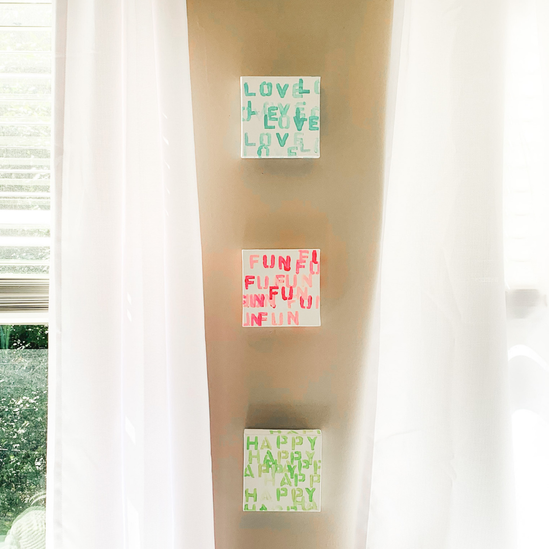 3 mini canvas pantings hung on a tan wall. The words love is stamped repeatedly on one in blue, the fun in pink on the middle, and the word happy in green on the last