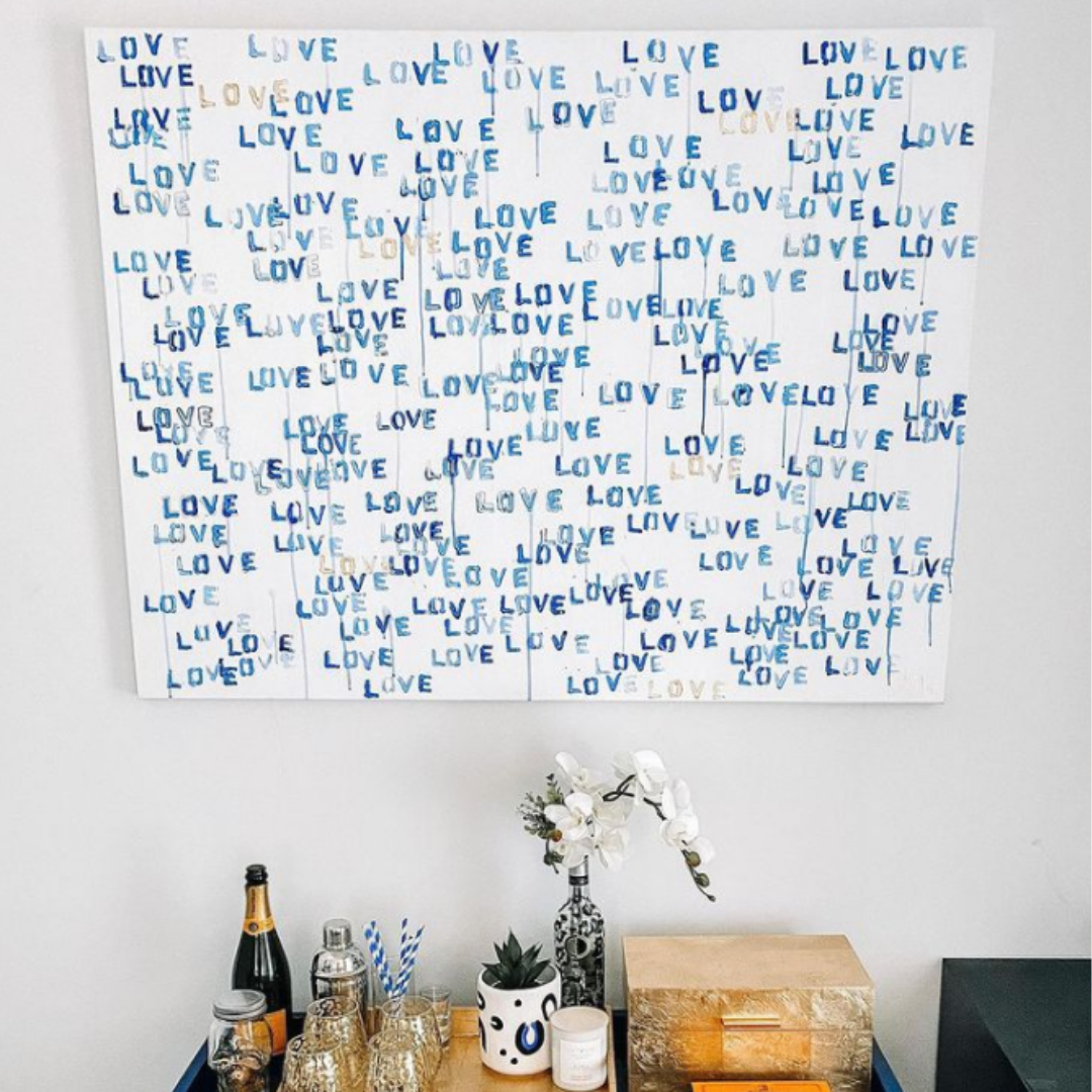 A large white canvas with the word love repeatedly stamped all over in different shades of blue. Hangs above a matching blue bar cart.