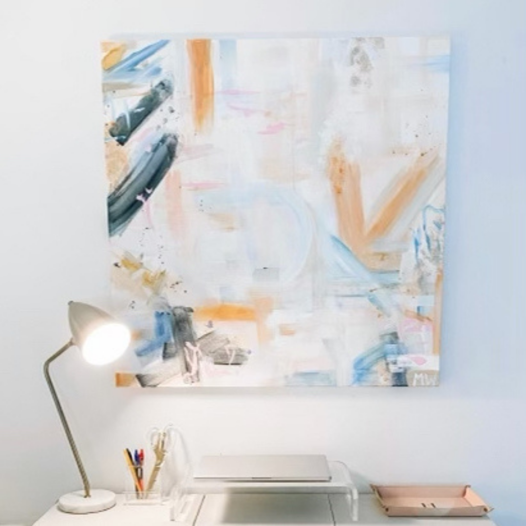 An abstract canvas painting with paint strokes of cream, orange, tan, and blue, hangs above a clean desk.