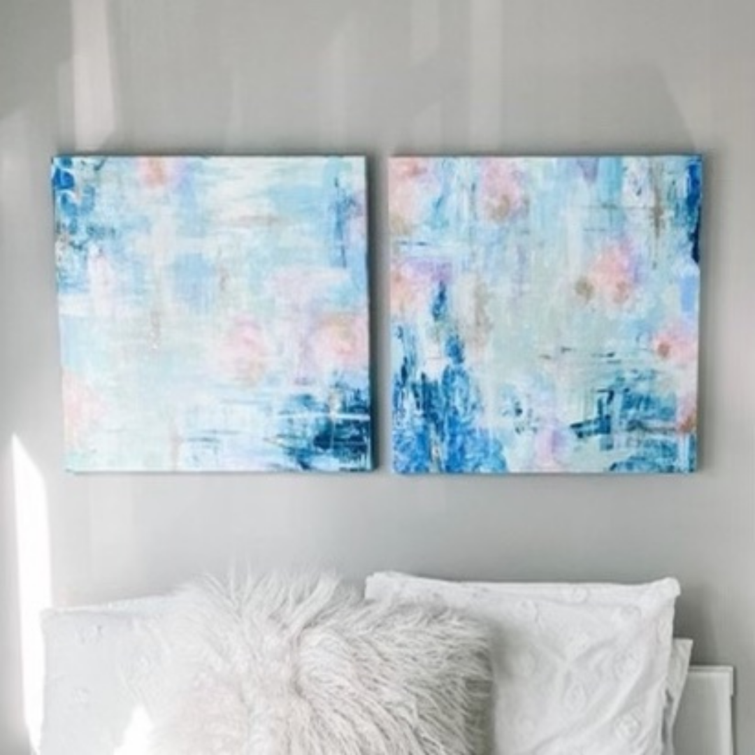 Two large canvases hang above a white bed. The paintings are abstract with minimal brush strokes mostly in blues. Color is concentrated in the corners with darker blues and small bits of orange and pink peak through the light blues.